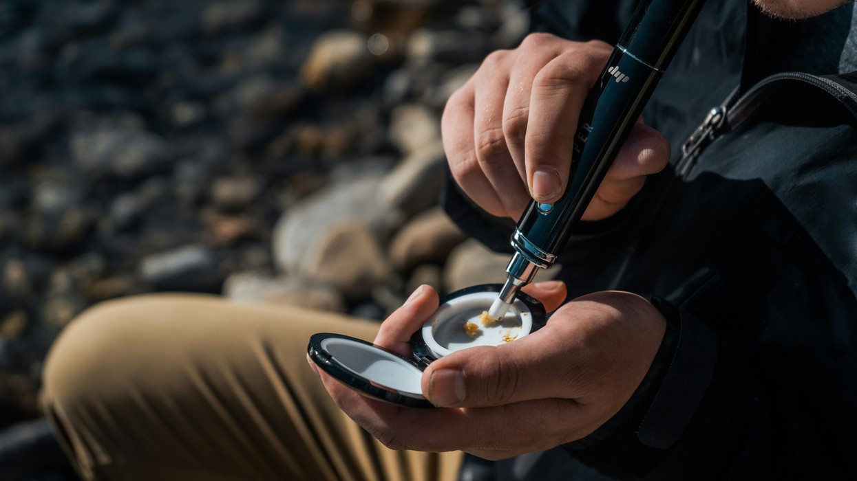 A man sits on a rocky slope, using a black device to consume cannabis resin.